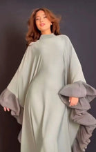 Woman wearing a sage green flowy abaya with layered grey ruffle sleeves, standing against a dark background